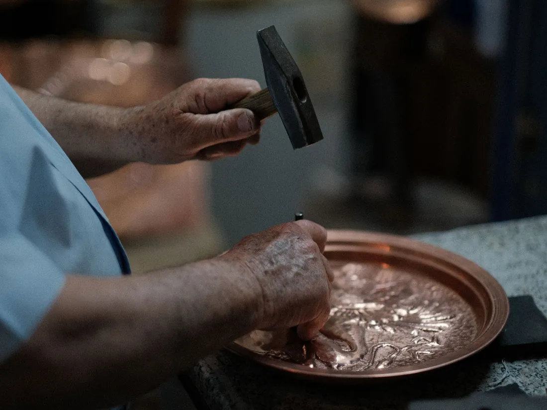 Manos sosteniendo un cincel y un martillo sobre una bandeja de cobre durante el trabajo de relieve